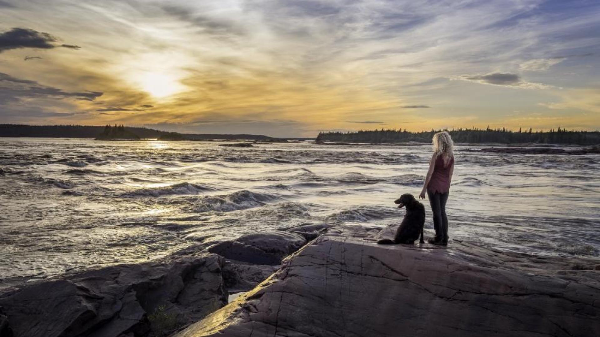 Woman and dog standing on rocks near the waters. They both are looking out to the horizon as the sun is setting in the background.